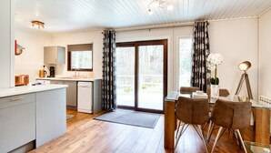 A modern kitchen with a central island, a dining area with a glass table, and a view through the sliding glass doors. - Otter With Mountain View and Enclosed Decking (Crianlarich)