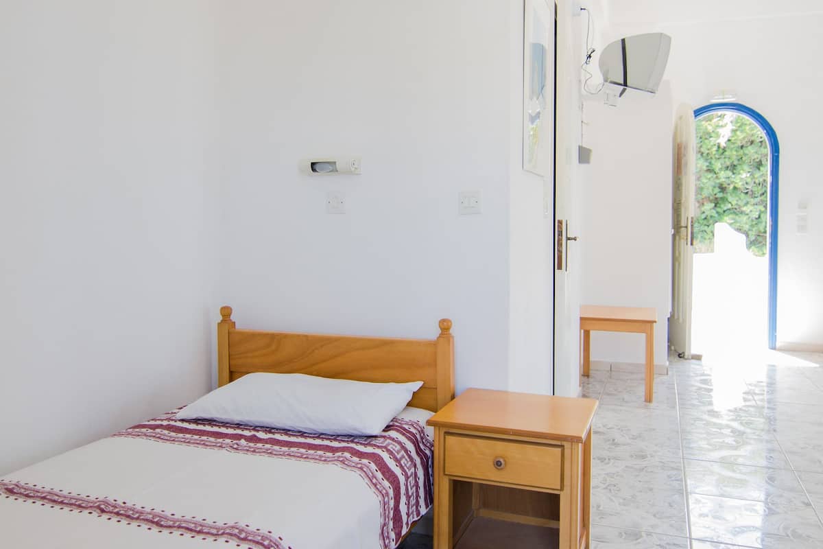 A small, clean room with a single bed, a wooden nightstand, and a small table. There is a door leading to an outdoor area.