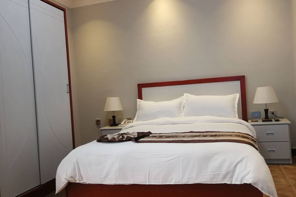 A neatly made bed with white linens, two bedside lamps, a nightstand, and a red-framed headboard.