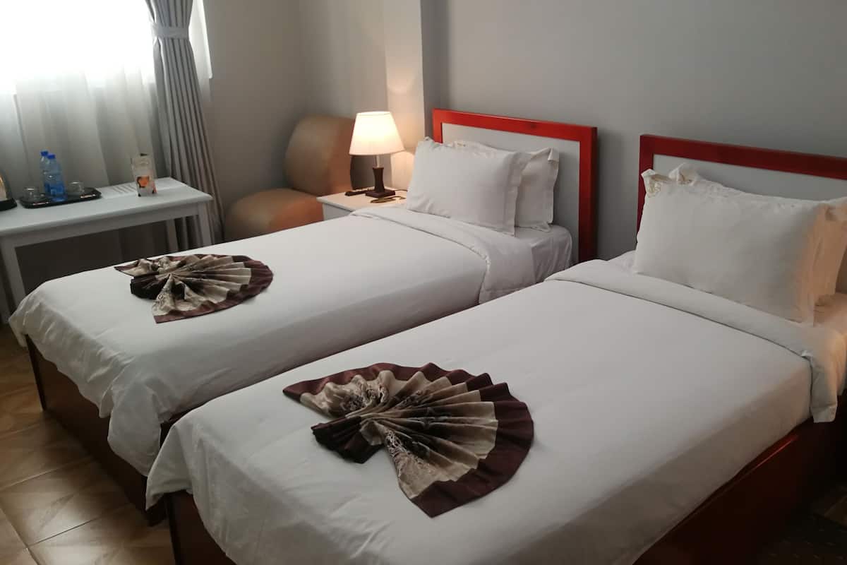 A hotel room with two beds, each with white linens and a decorative fan on the bed. There is a nightstand with a lamp and a phone, and a window with curtains.