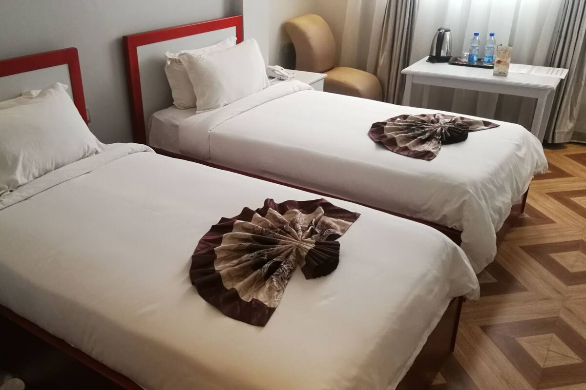 Two beds with white linens and red headboards, a bedside table with water bottles, and a chair.