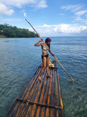 A person standing on a bamboo raft in clear water with a forested island in the background.