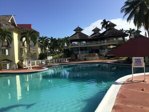 A swimming pool with a poolside area, palm trees, and a multi-story building with a red roof.