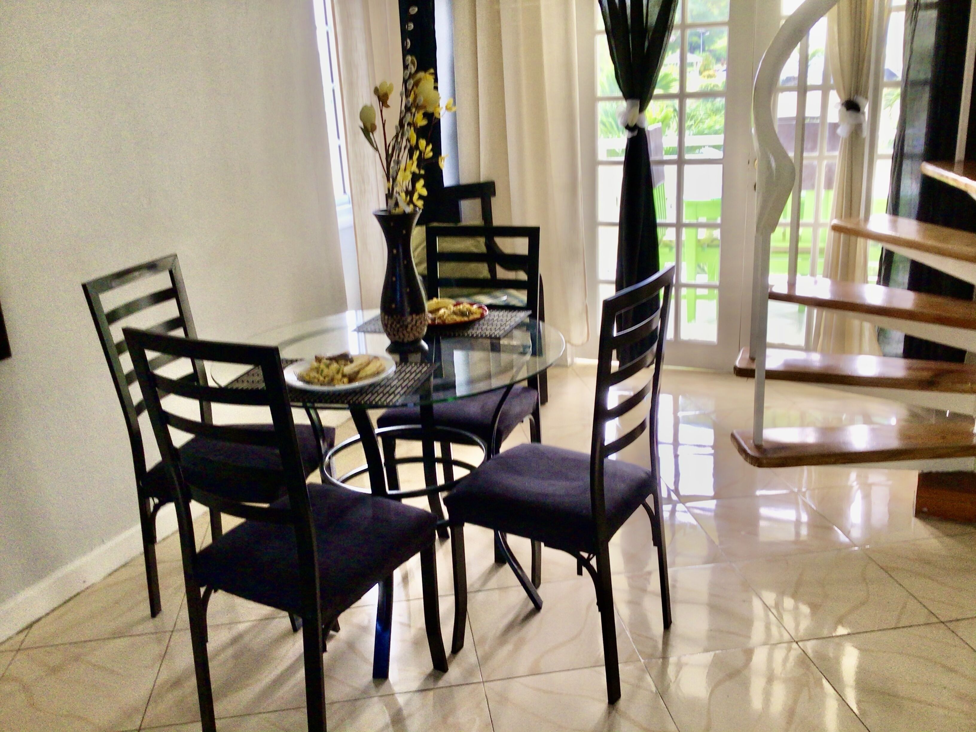 A dining area with a glass table, four chairs, a vase with flowers, and a view of a garden through a window.