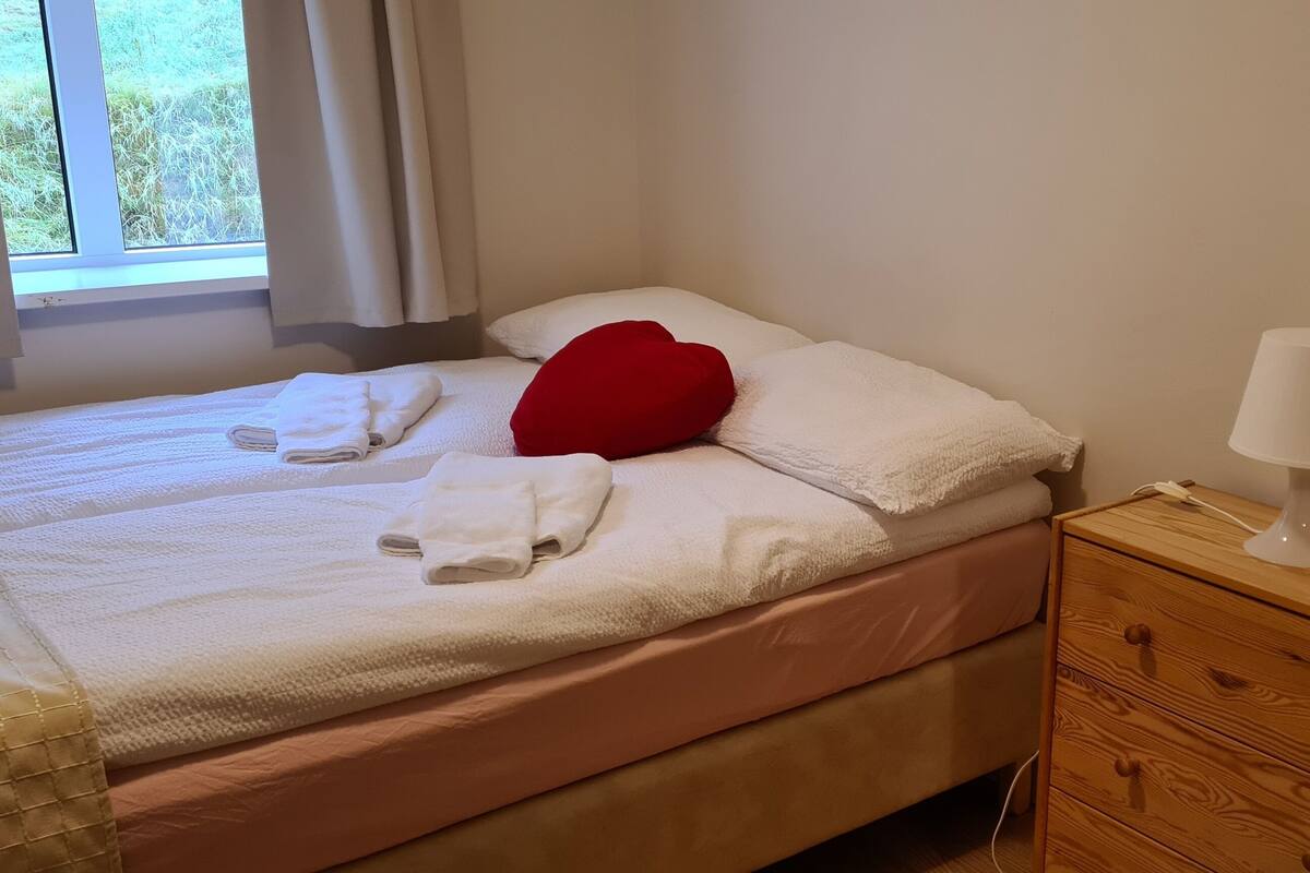 Un lit bien fait, avec du linge de lit blanc et un coussin rouge, une lampe de chevet et une table de chevet en bois dotée de tiroirs.