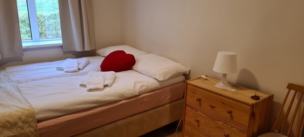 A neatly made bed with white linens and a red pillow, a bedside lamp, and a wooden nightstand with drawers. - Guesthouse Stekkatún (Höfn)
