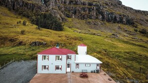 A two-story building with a red roof and white walls, situated on a grassy hillside with a rocky cliff in the background.