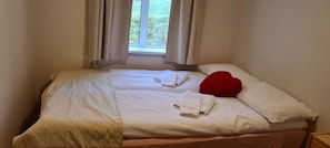 A neatly made bed with two towels and a red pillow, a window with curtains, and a view of greenery outside.