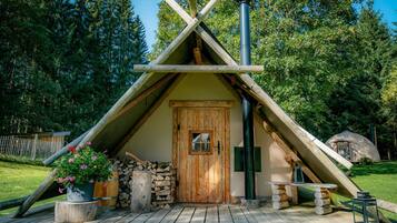 Una cabaña de madera tipo A con puerta, chimenea y un pequeño porche con una maceta y leña.