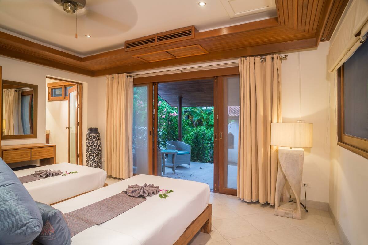 A hotel room with a double bed, a bedside table, a lamp, a chair, and a view of the garden through the sliding glass door.