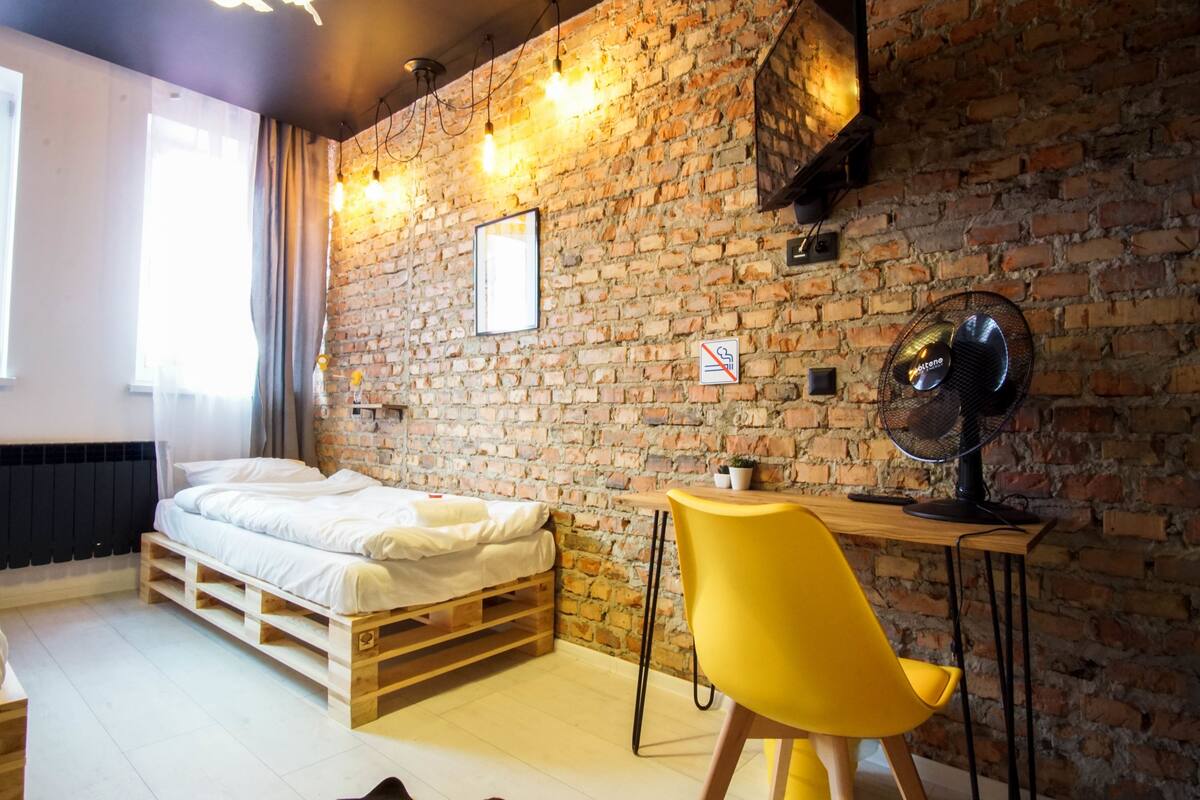 A room with a brick wall, a bed, a yellow chair, and a desk with a fan.