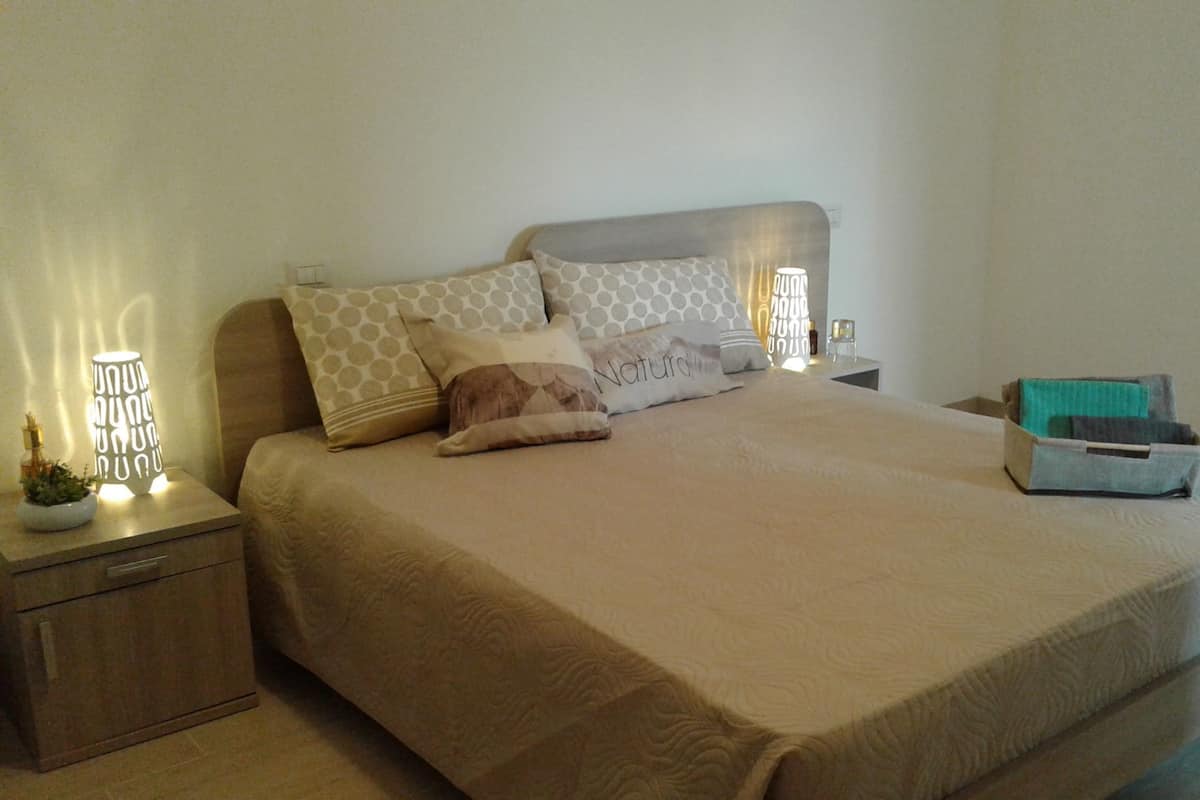 Una cama bien hecha con edredón color beige, varios almohadones y una mesita de noche con lámpara y una pequeña planta.