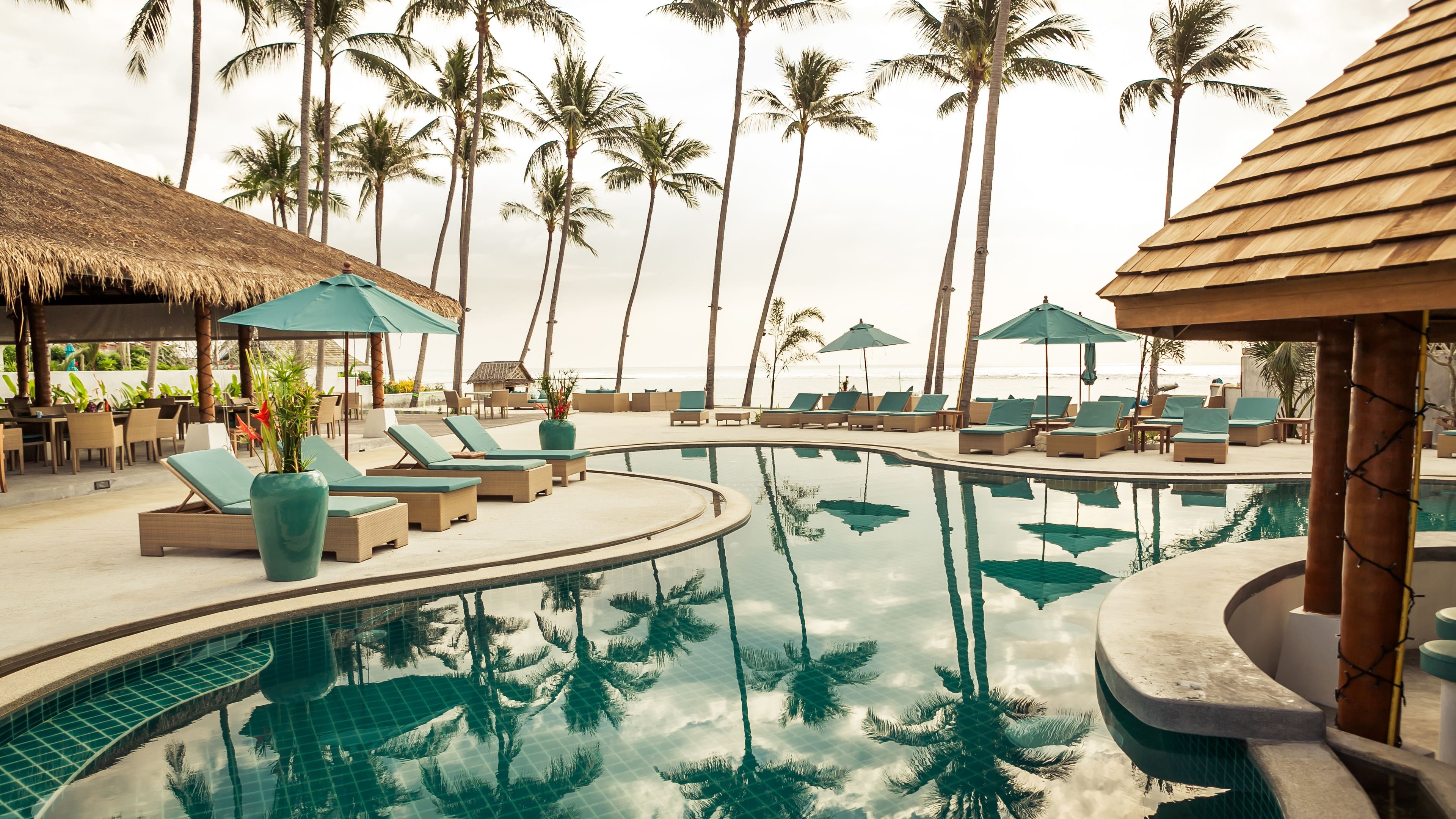 A poolside area with lounge chairs and umbrellas, surrounded by palm trees and a view of the beach.