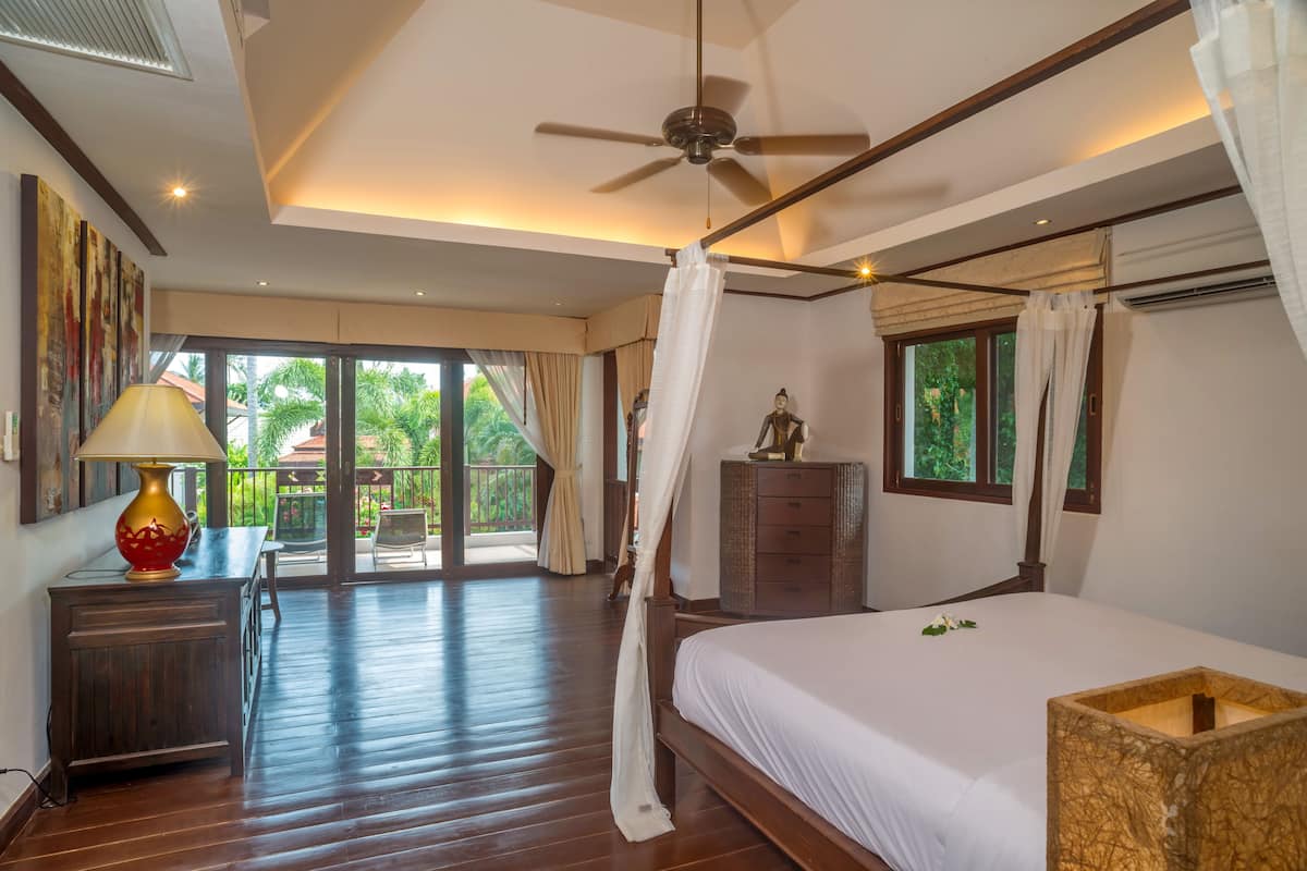 A bedroom with a bed, a ceiling fan, a nightstand, a lamp, and a view of the outdoors.