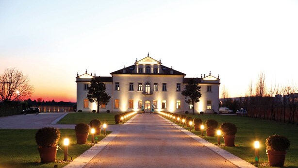 Un edificio ampio e simmetrico con un ingresso centrale, circondato da un vialetto ben illuminato e piante in vasi disposti regolarmente.