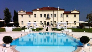 Una grande piscina rettangolare con superficie d'acqua blu cristallina, circondata da un solarium in pietra. Sullo sfondo, un edificio a due piani con tetto in tegole rosse e numerose finestre.
