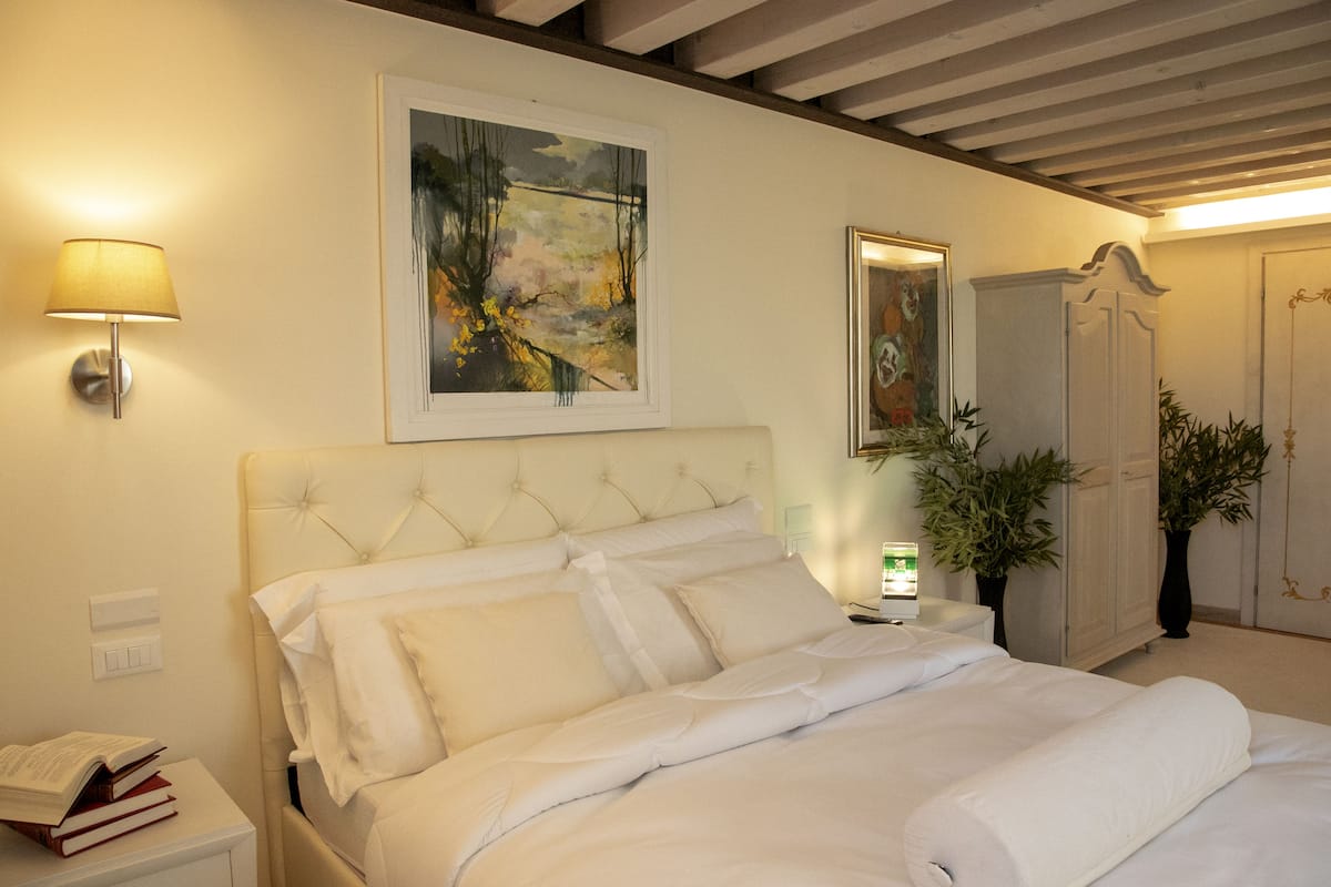 Una cama bien tendida con sábanas blancas, una lámpara de noche, un cuadro enmarcado sobre la cama y un armario con espejo.