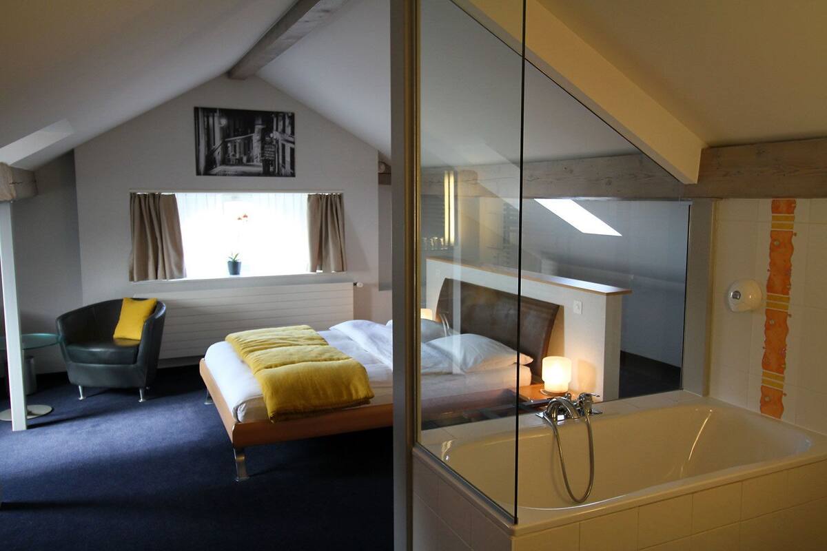Quarto com cama, cadeira, janela com cortinas, radiador e banheira.