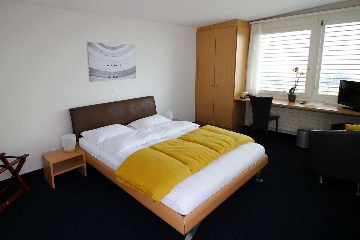 Quarto de hotel com cama, escrivaninha com cadeira, guarda-roupa e janela com persianas.