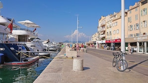 A marina with boats docked, a paved walkway, and buildings with shops. - Villa Colibri (Lorgues)