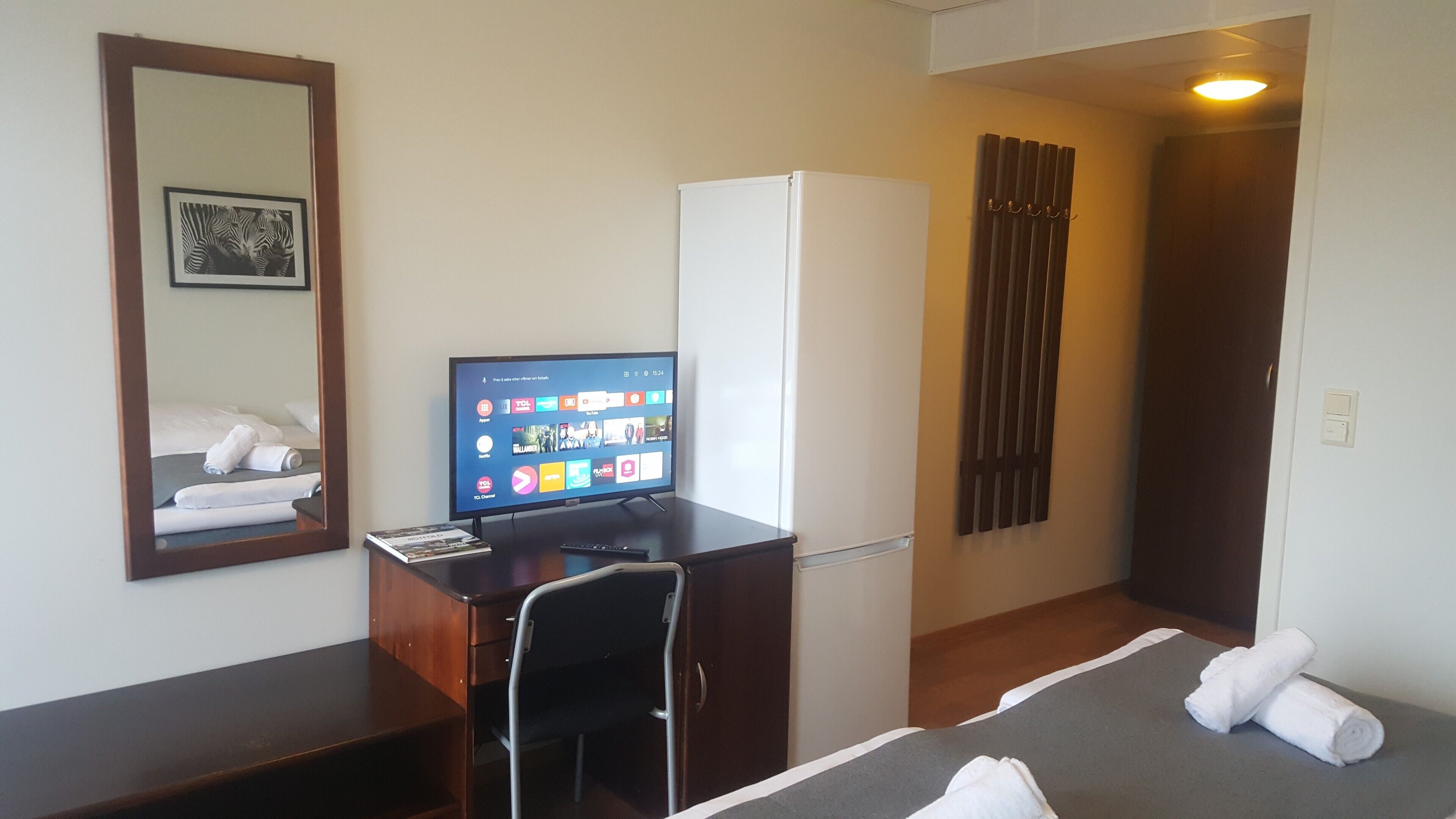 Habitación de hotel con escritorio, televisor mostrando varios canales, cama con ropa de cama impecable, espejo y puerta que da a otra habitación.