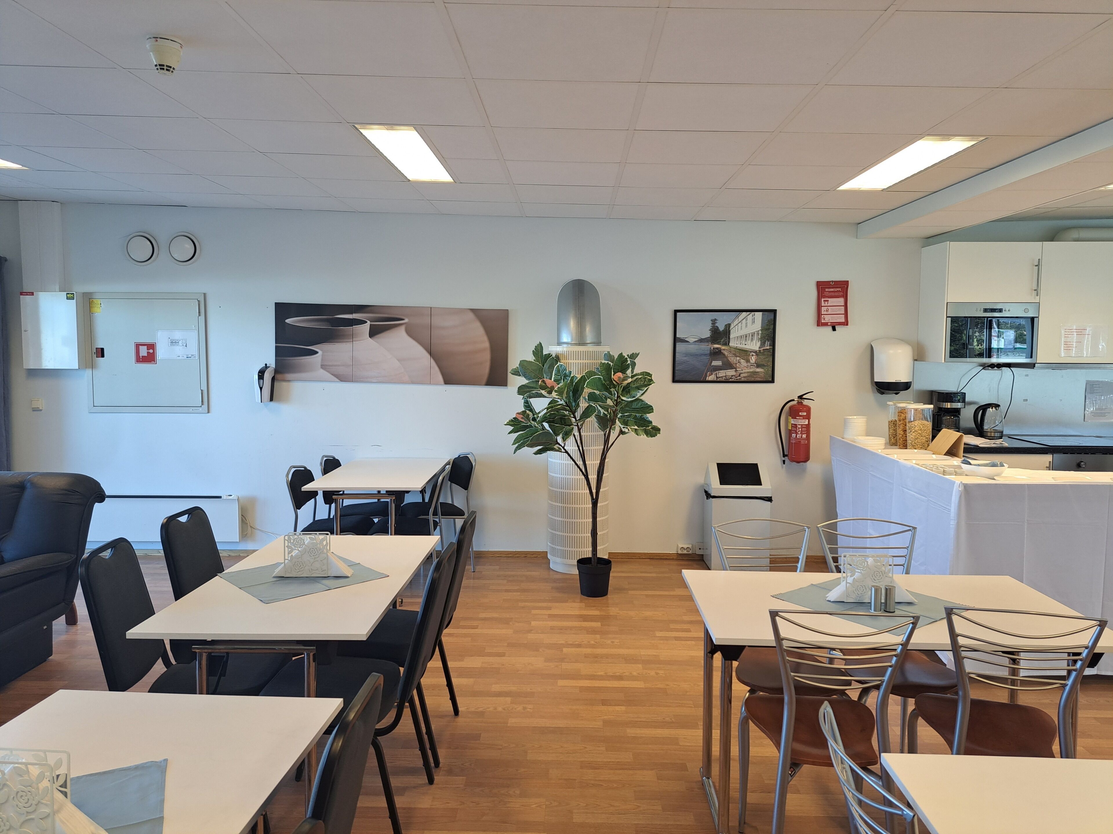A modern dining area with a central plant, a kitchen area with a coffee machine, and a fire extinguisher on the wall.