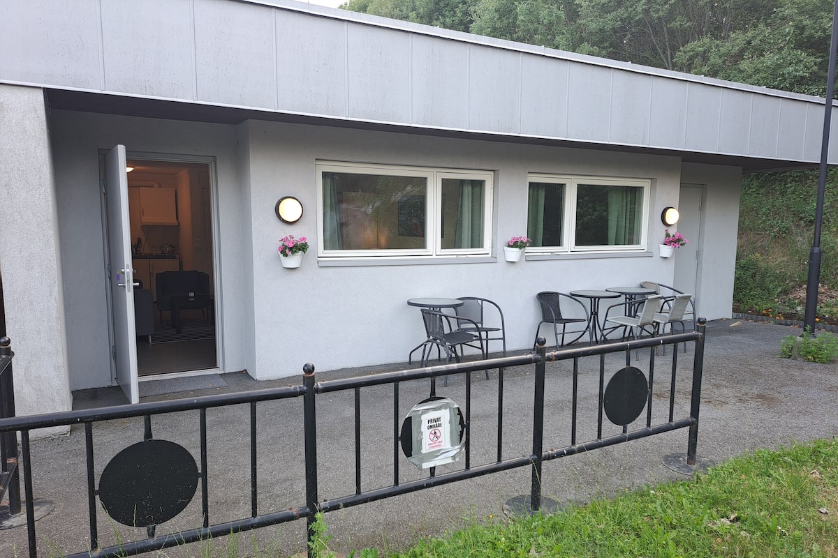 A building with a white exterior, a covered outdoor seating area with tables and chairs, and a small entrance with a door.