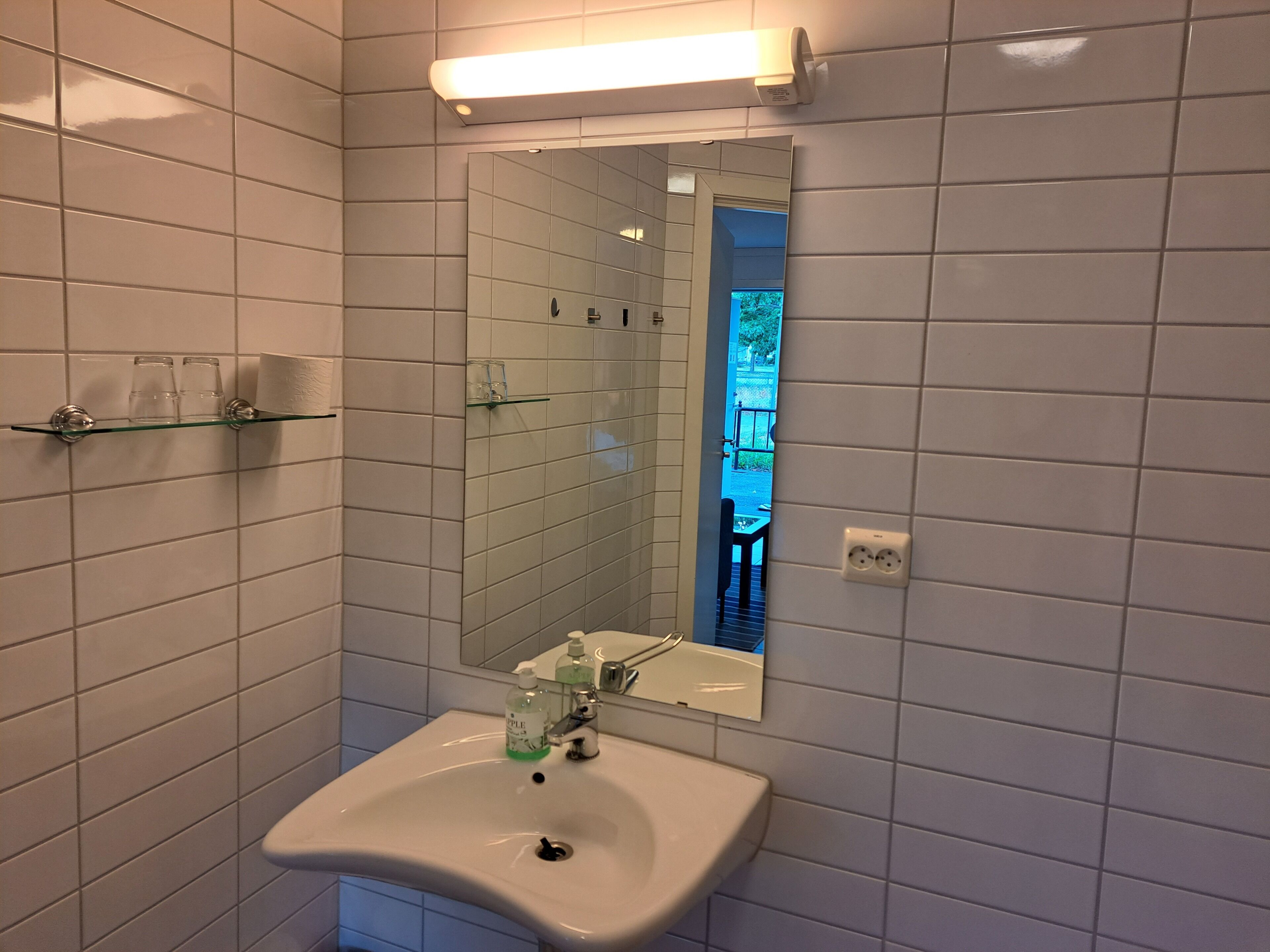 Ett badrum med vita kakelväggar, en hylla i glas med toalettartiklar, ett handfat med en spegel ovanför och en synlig utomhusyta som återspeglas i spegeln.