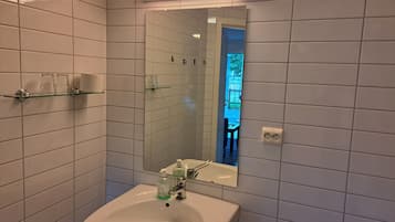 Ett badrum med vita kakelväggar, en hylla i glas med toalettartiklar, ett handfat med en spegel ovanför och en synlig utomhusyta som återspeglas i spegeln.