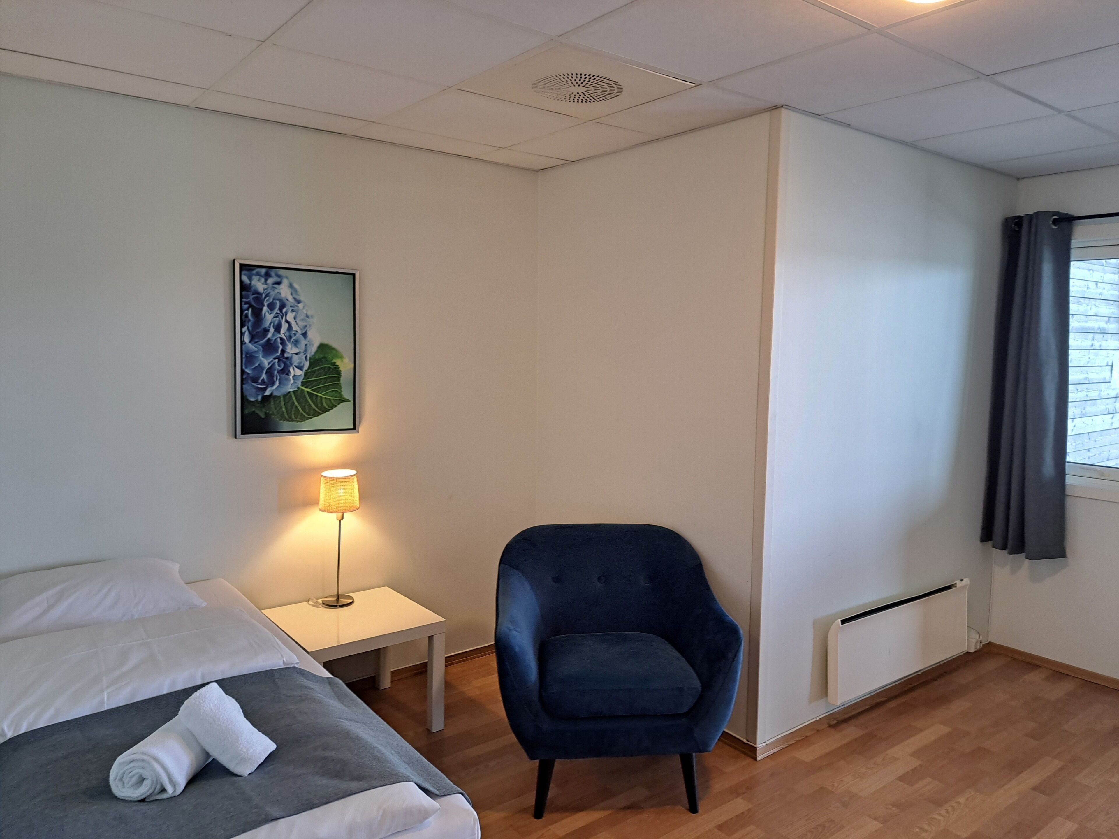 Ett hotellrum med en säng, en stol, ett litet bord med en lampa och ett fönster med gardiner.