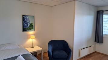 Ett hotellrum med en säng, en stol, ett litet bord med en lampa och ett fönster med gardiner.