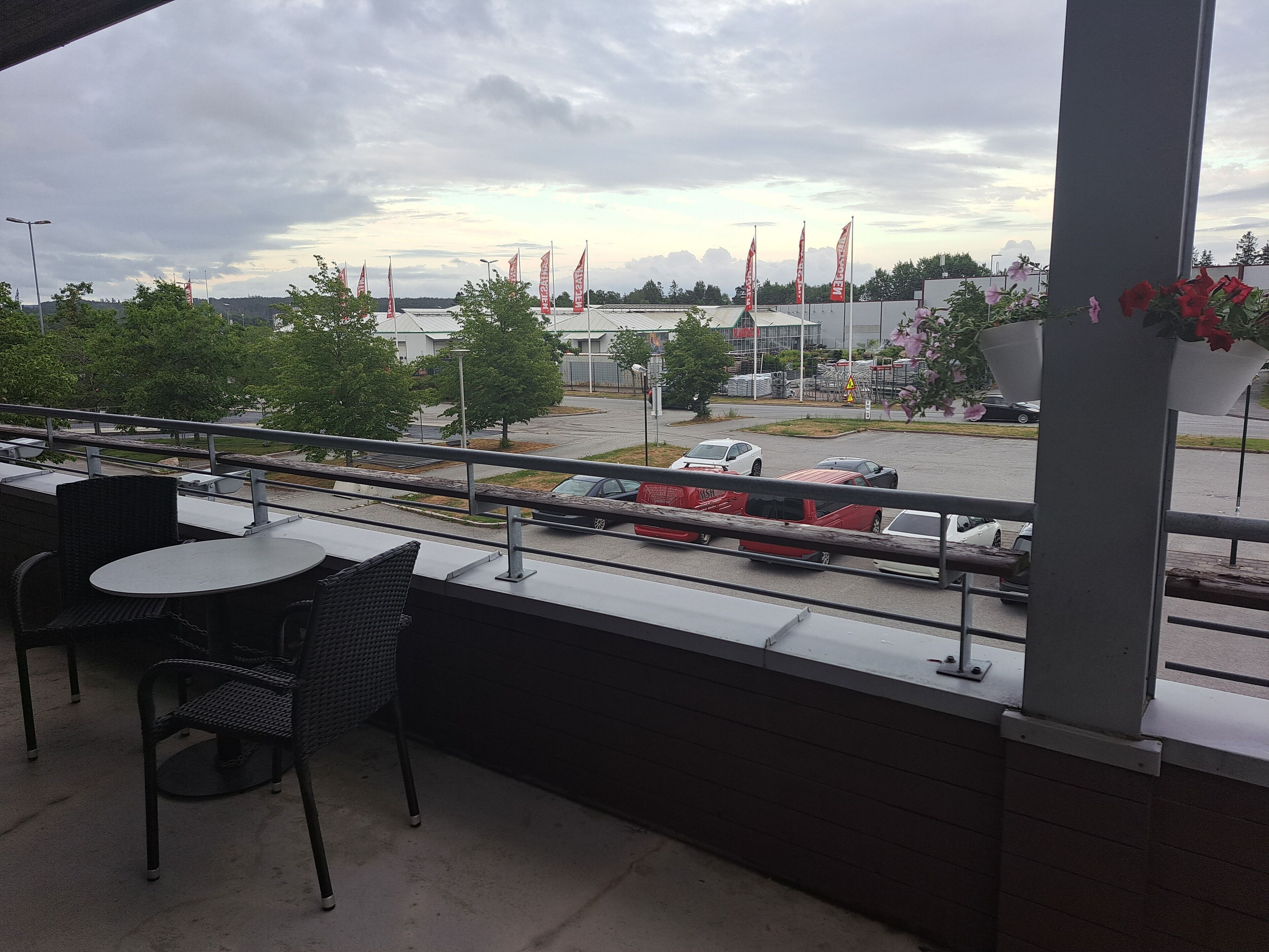 Un balcón con mobiliario exterior, vista a un estacionamiento y un edificio con banderas al fondo.