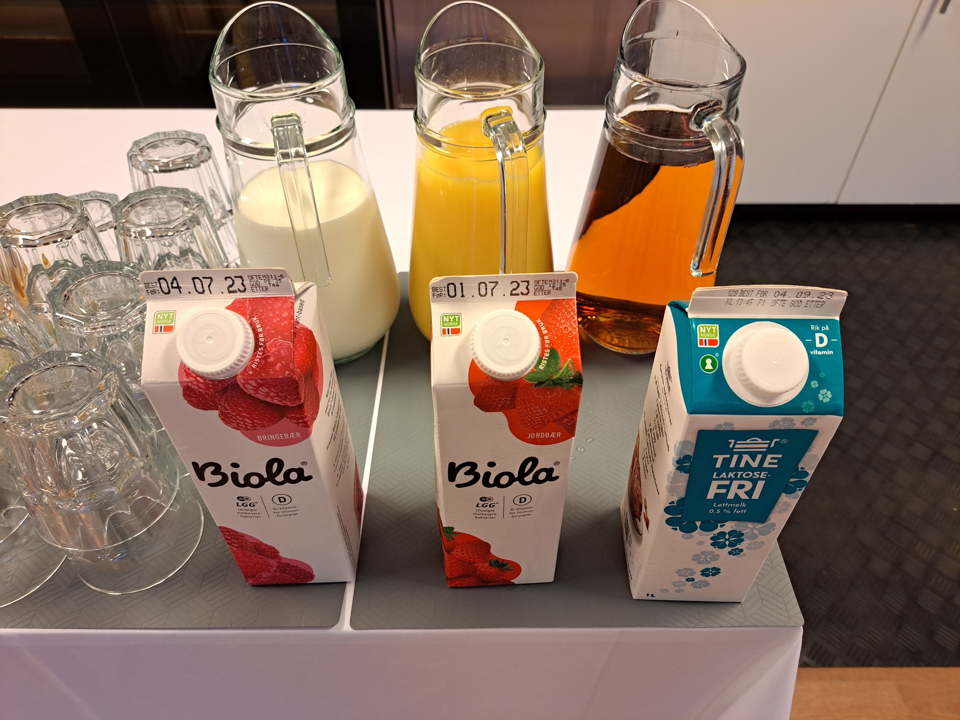 Tres cartones de jugo Biola, un vaso de leche y un vaso de jugo de naranja sobre una mesa.