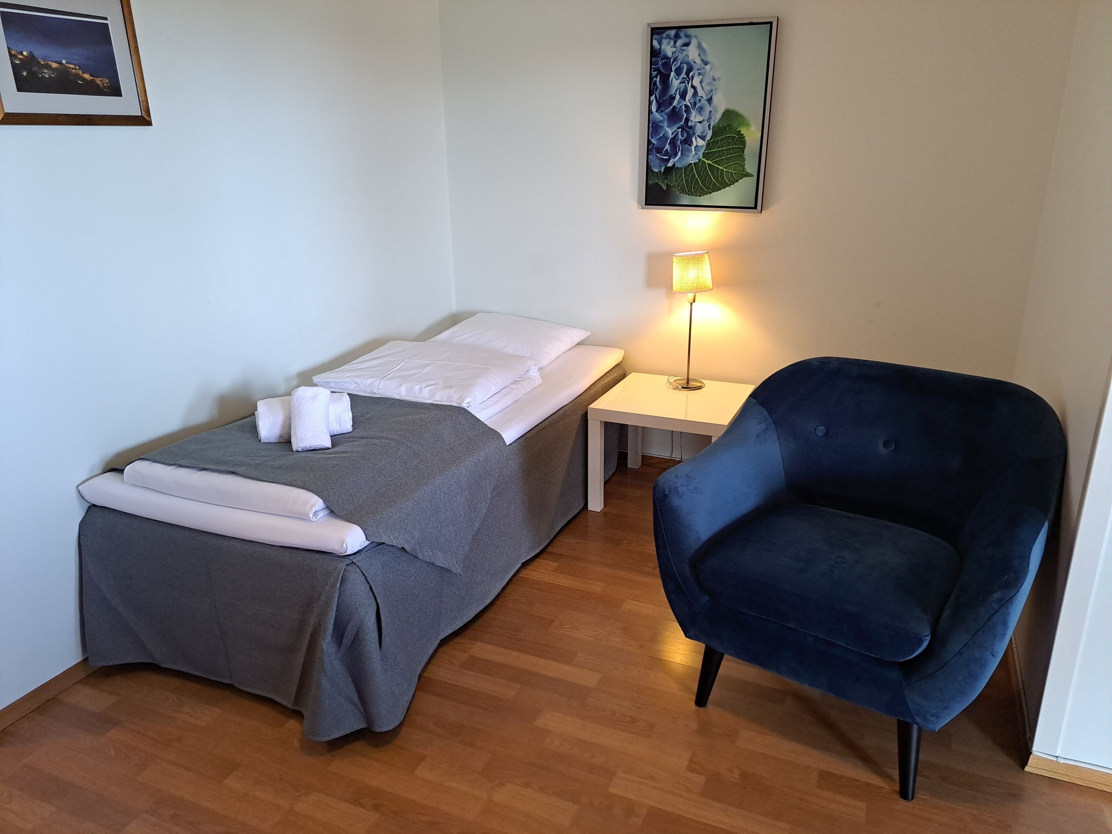 Habitación de hotel con cama, una silla, mesita de noche con lámpara y un cuadro en la pared.