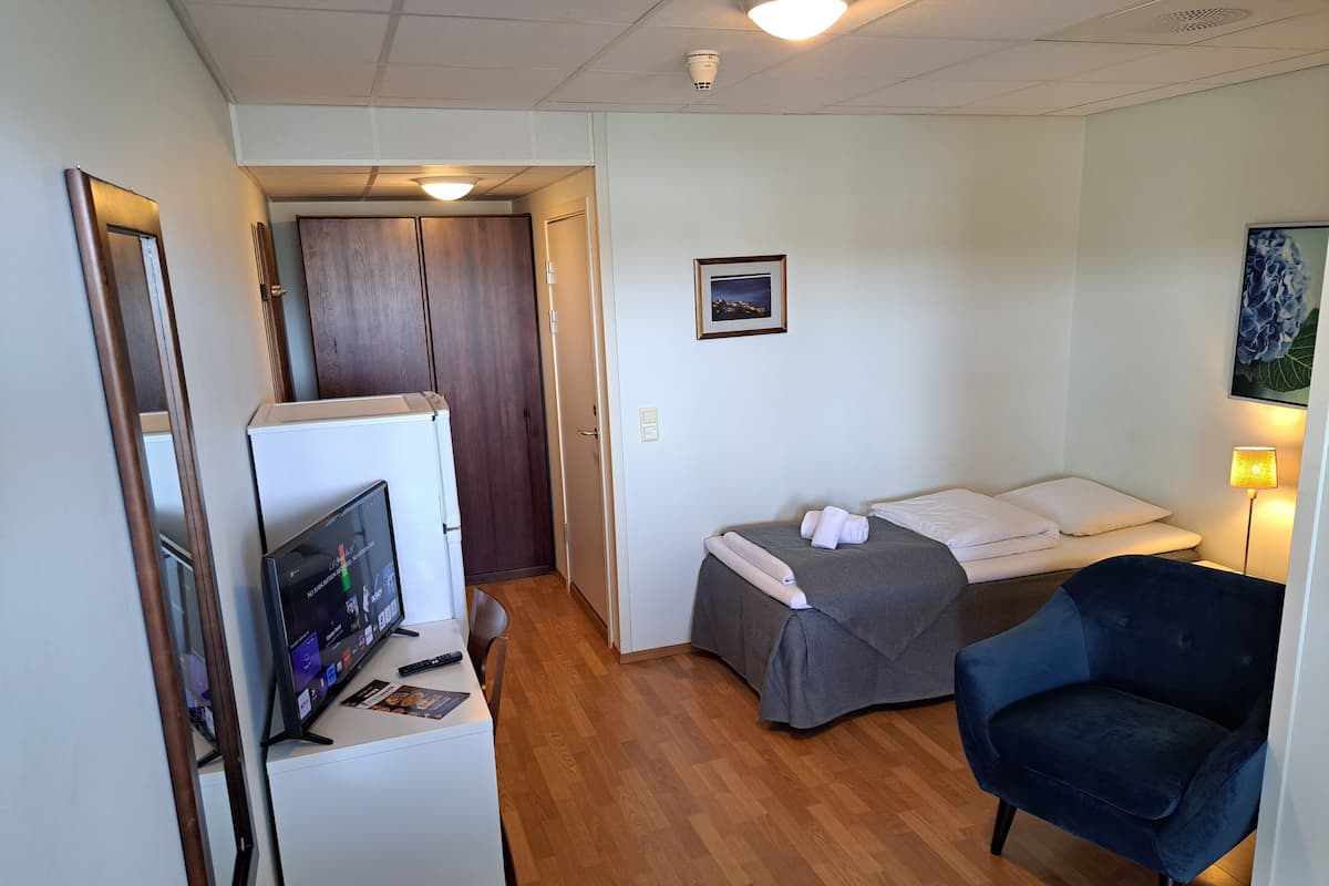 A small, well-lit room with a bed, a chair, a desk with a computer, and a door leading to another room.