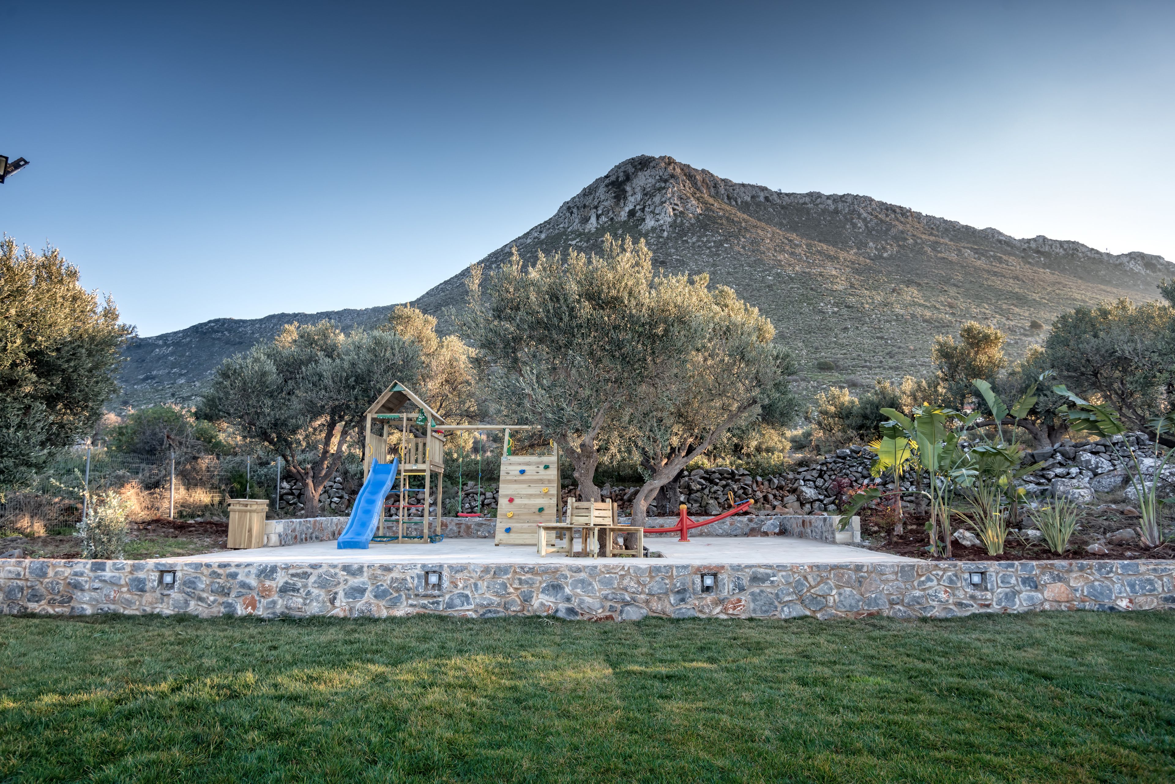 Un parque infantil con paredes para escalar y un tobogán, rodeado de un muro de piedra y árboles, con una montaña al fondo.
