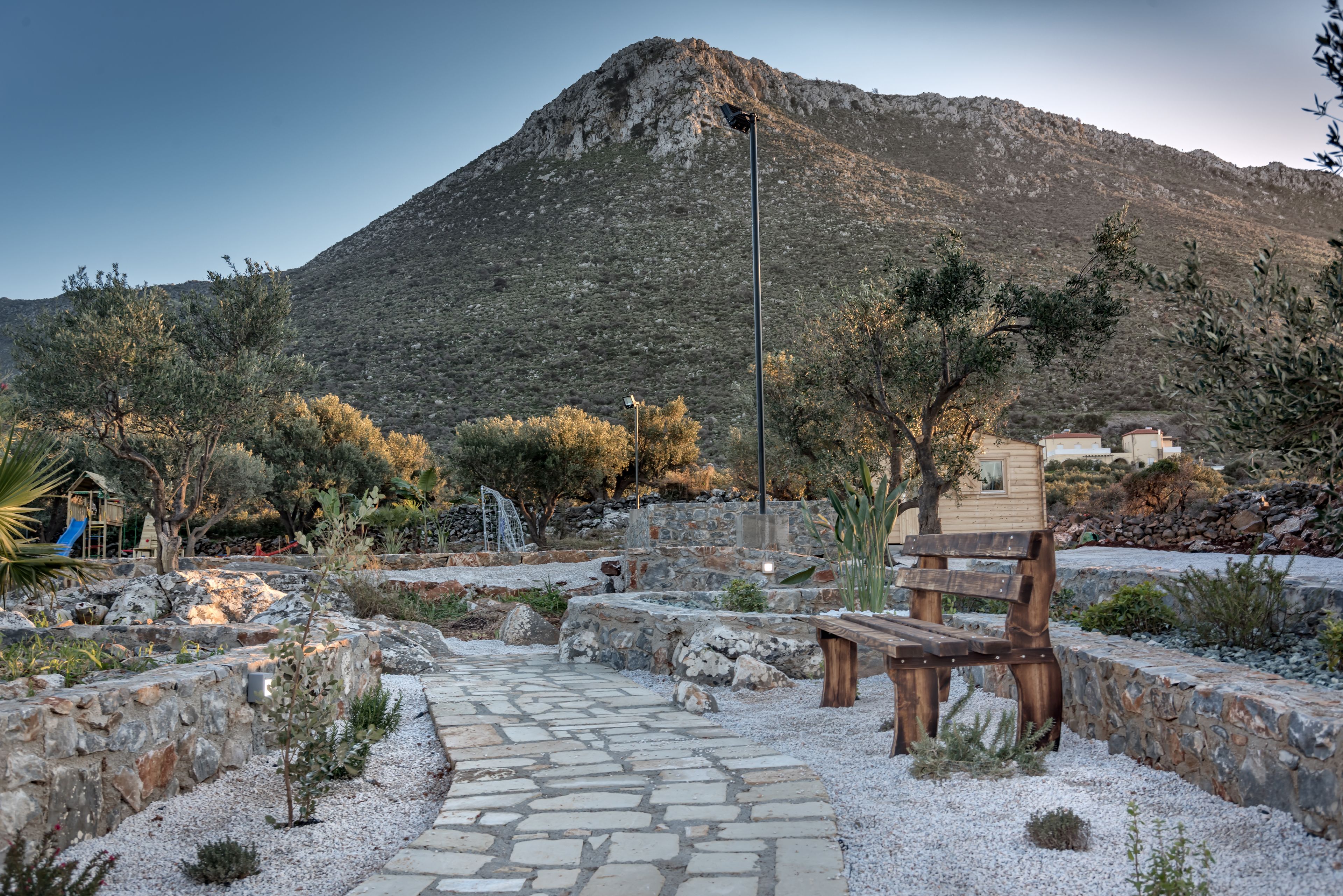 A stone pathway leading to a wooden bench and a playground area with a mountain in the background.