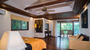 A hotel room with a bed, a sofa, a ceiling fan, and a large window with a view of greenery. - Baan Tawan (Koh Samui)