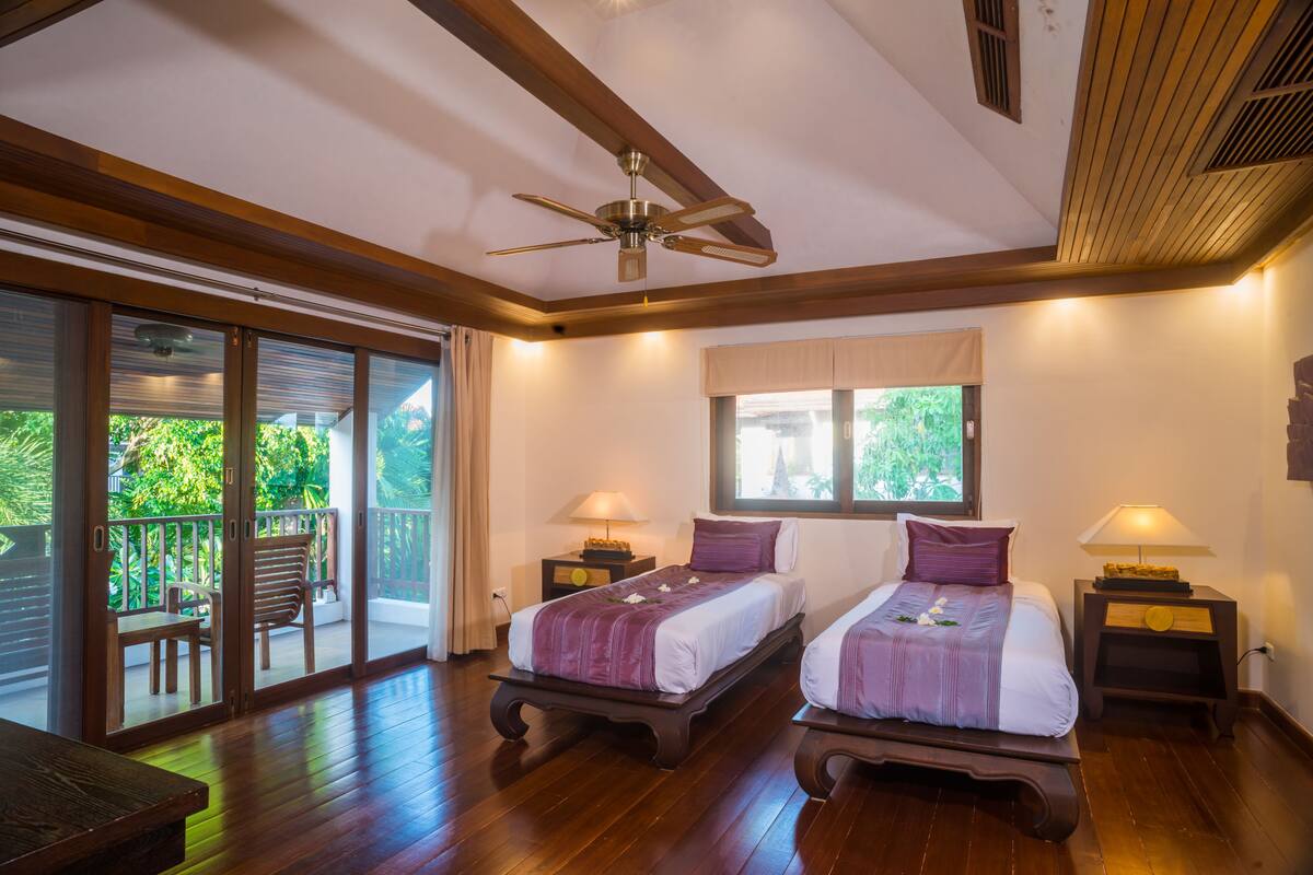 Una habitación con dos camas, ventilador de techo, balcón con mesa y sillas, y ventana con vistas a la vegetación.