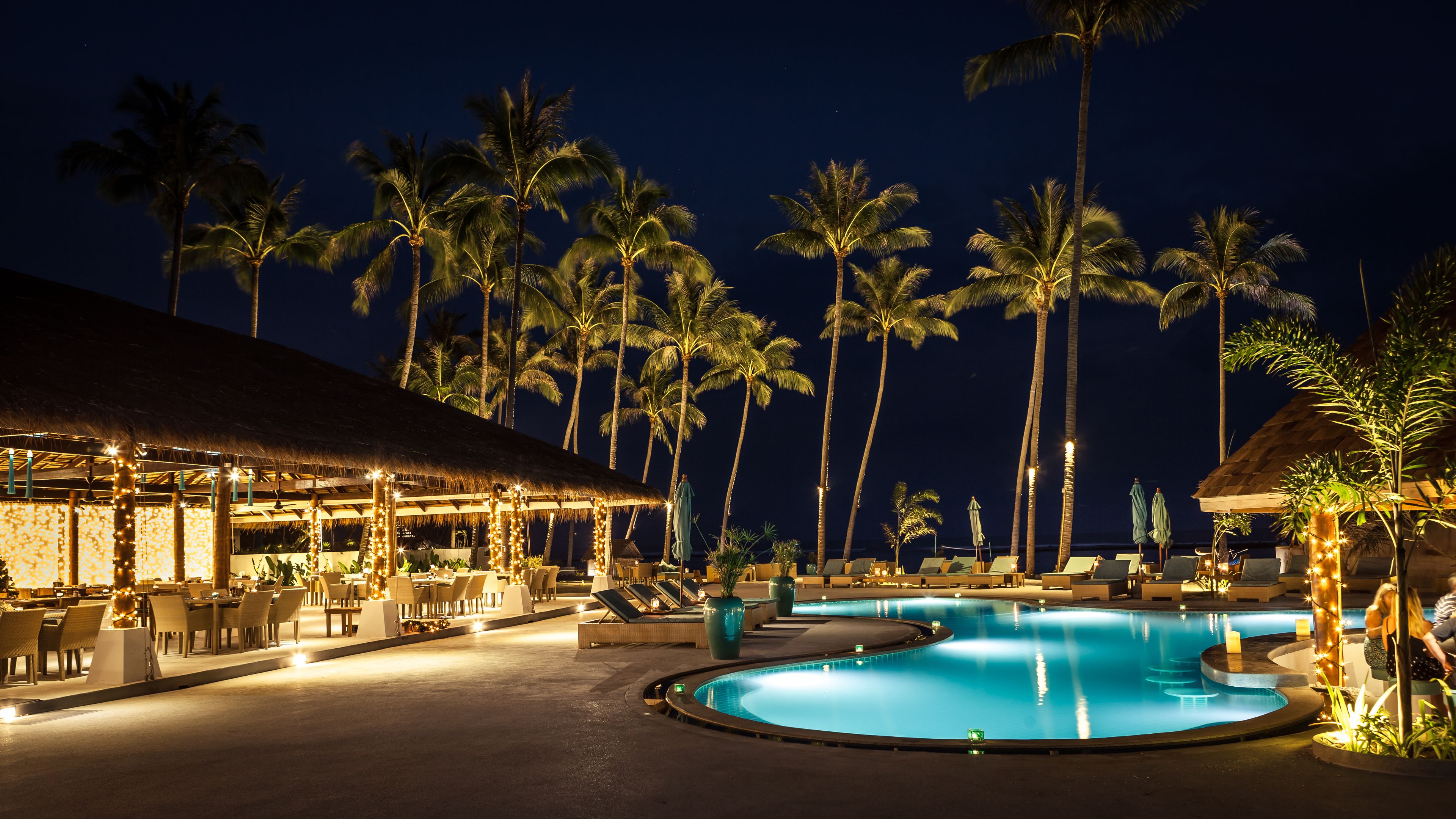 Un espace piscine dans un complexe hôtelier, avec des chaises longues, un bar et des palmiers, le tout illuminé la nuit.