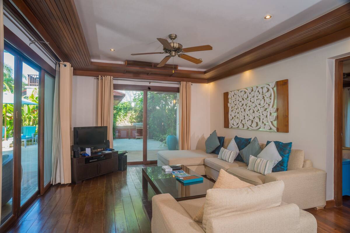 Amplia sala de estar con ventilador de techo, televisor de pantalla plana, sofá con cojines, mesa de centro y puerta corrediza de vidrio que da a un patio al aire libre.