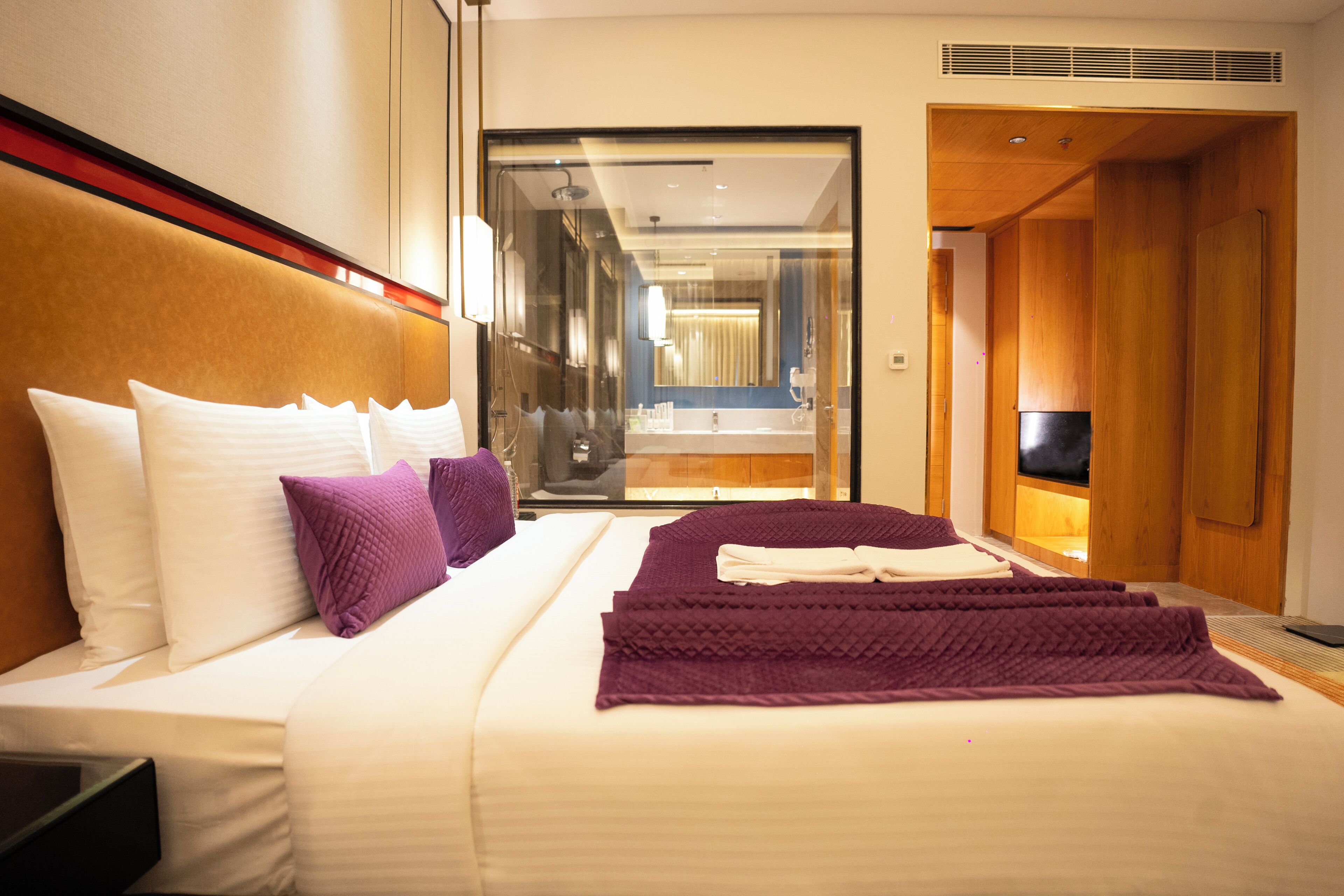 A hotel room with a bed, pillows, a blanket, a TV, and a bathroom visible through a glass door.