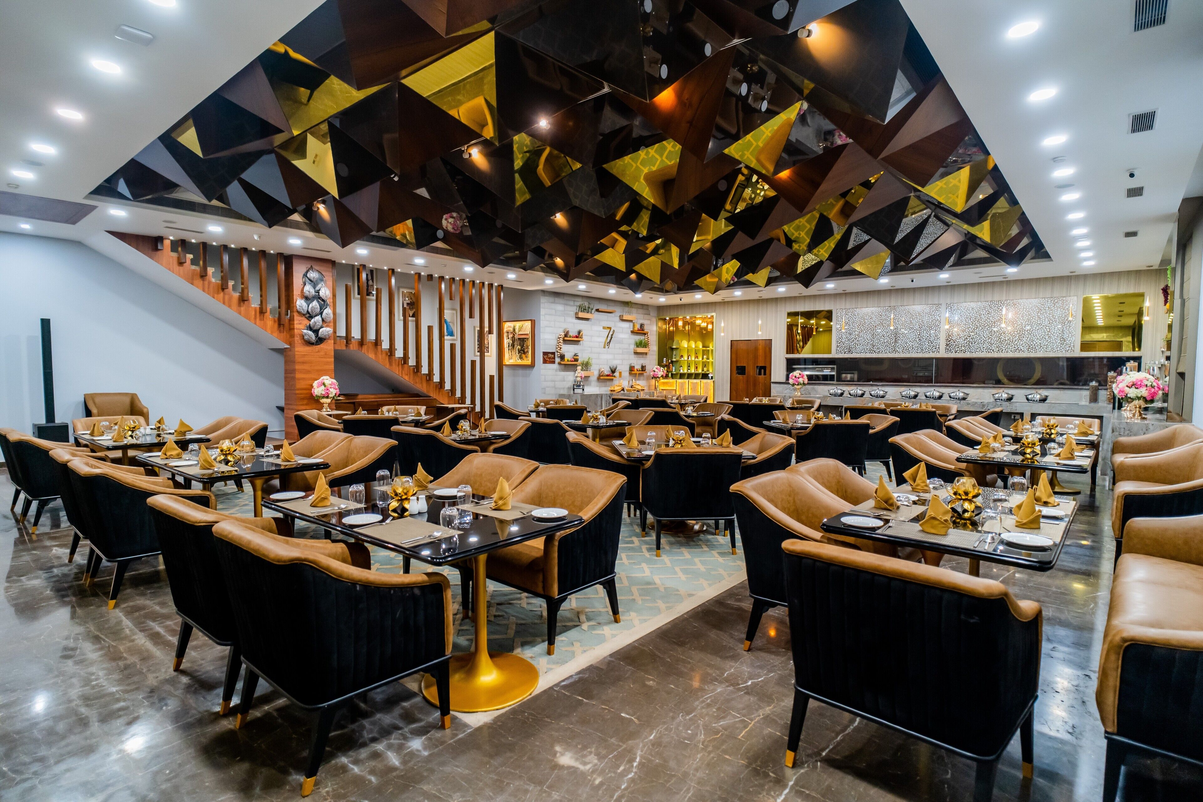 A modern restaurant with a geometric ceiling design, black and gold chairs, and a central staircase.