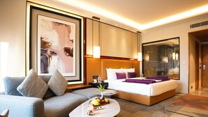 A modern hotel room with a sofa, a bed with a headboard, a coffee table with fruit, and a large framed artwork. - The Radiant Resort (Gorakhpur)