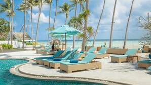 Un espace aménagé au bord de la piscine, avec des chaises longues et des parasols, des palmiers et une vue sur l’océan.