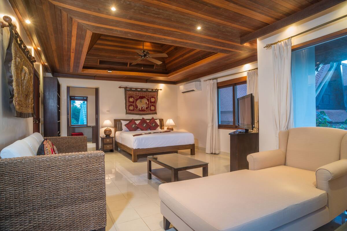 A hotel room with a wooden ceiling, a bed with red pillows, a sofa, a coffee table, and a TV.