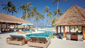 Un espace piscine aménagé dans un complexe hôtelier, avec des chaises longues, des parasols et une structure dotée d’un toit de chaume.