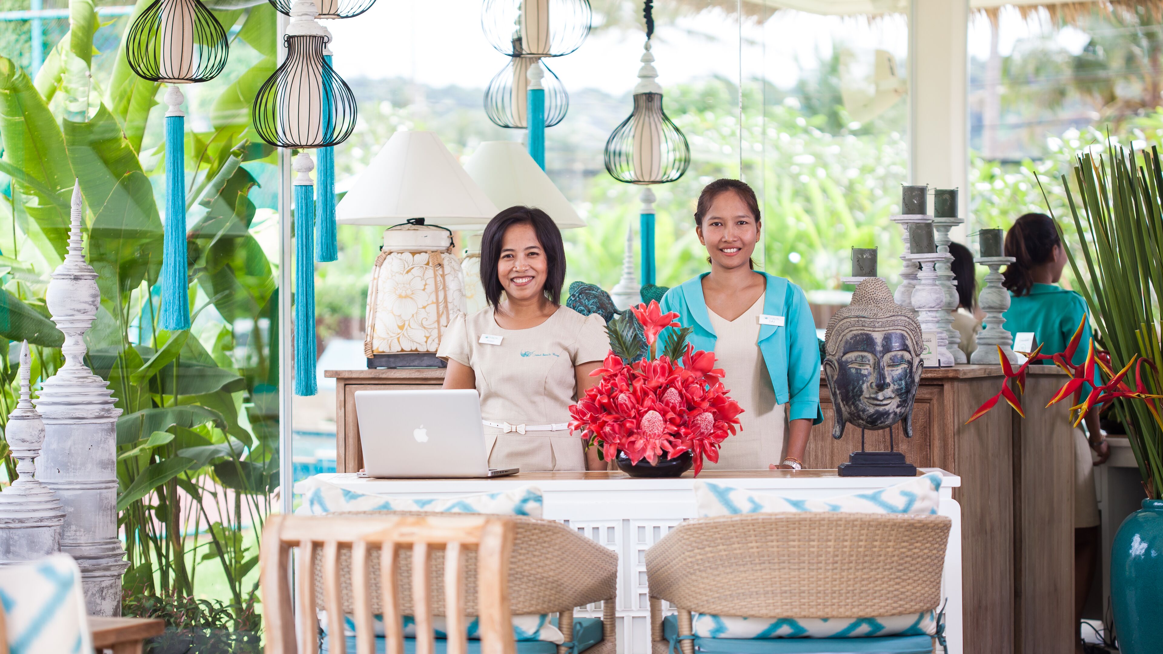 A reception desk with a laptop, a vase of red flowers, and a Buddha head sculpture.