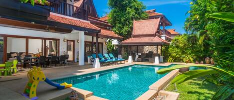 A swimming pool with a slide, surrounded by a garden and a building with a red-tiled roof.