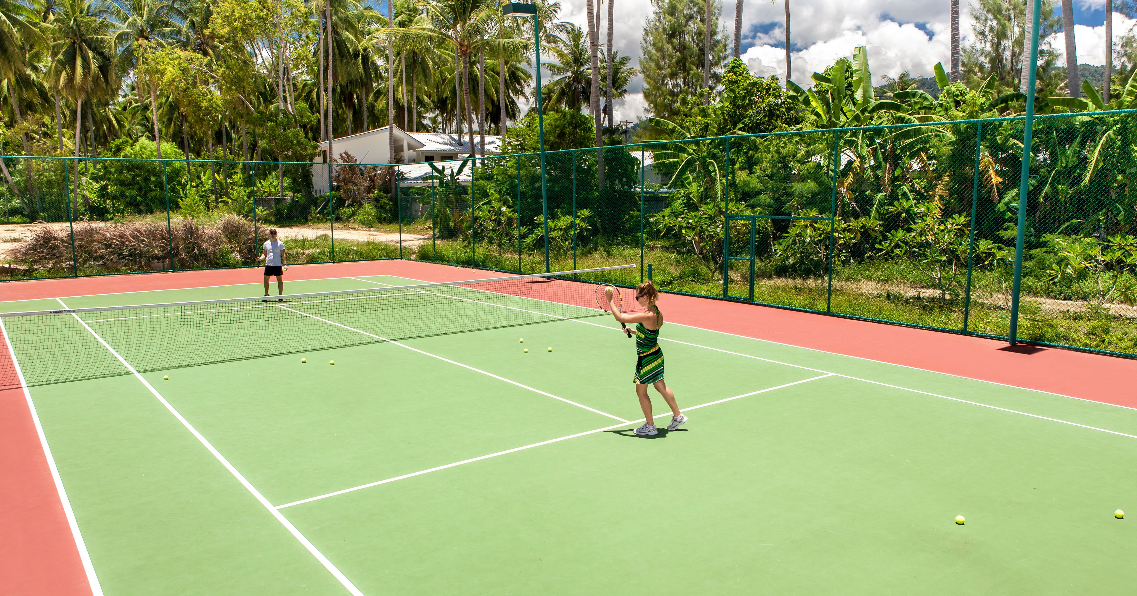 A tennis court with a net, surrounded by a green fence and palm trees.